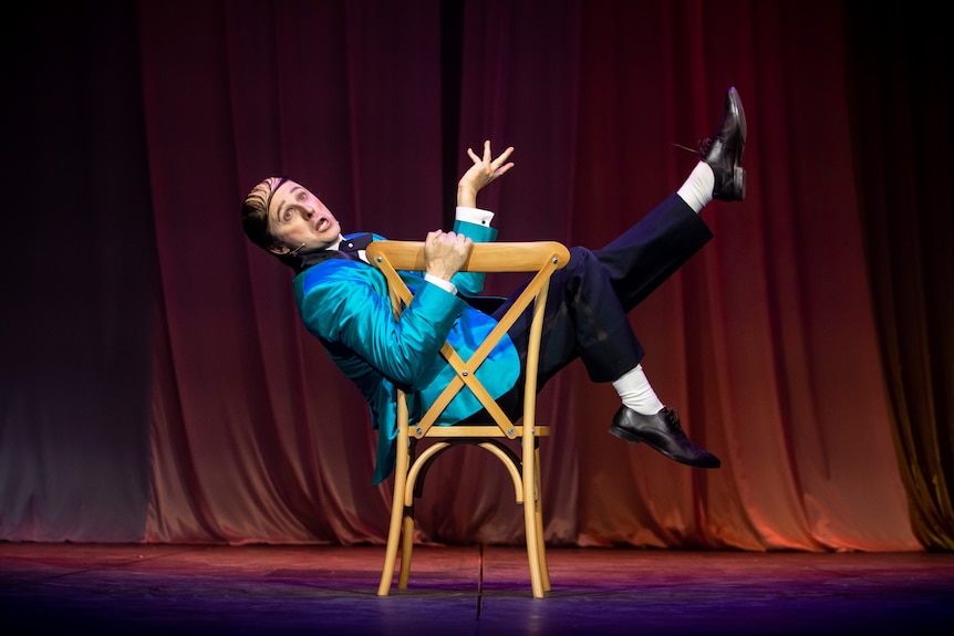 Matthew Whittet kicks his feet up from a chair onstage as Norman Gunston, in a shiny blue blazer with hair slicked in a combover