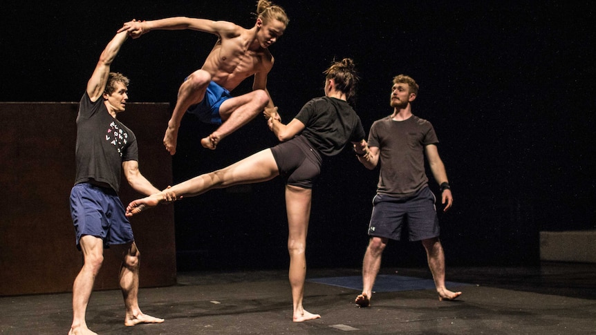 Acrobat Dylan Phillips dressed in shorts leaps over a fellow performer's leg.