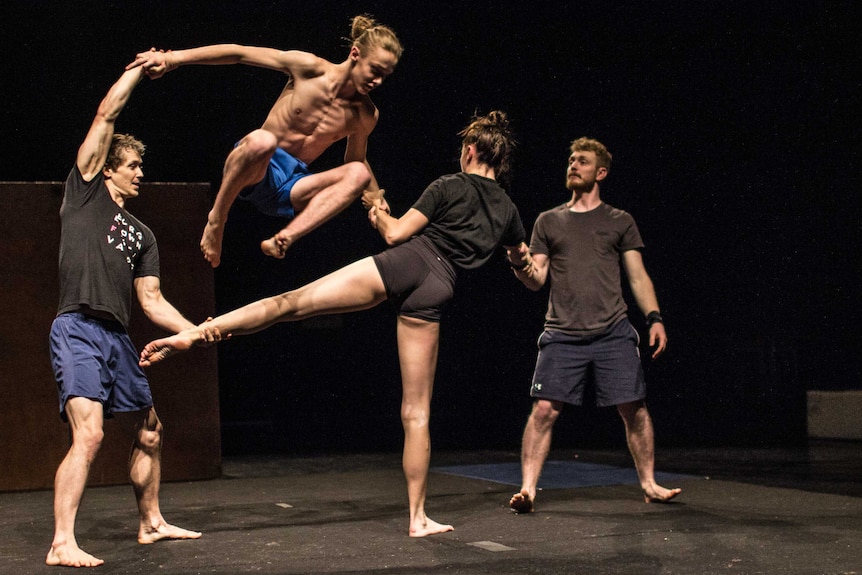 Acrobat Dylan Phillips dressed in shorts leaps over a fellow performer's leg.