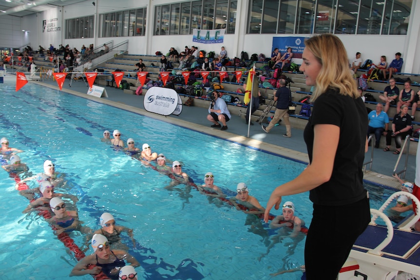 Olympic champion Ariarne Titmus stands on side of pool and talks to swimmers in pool.