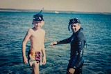 A young man in wetsuit and snorkelling gear stands in the water beside a young boy in swimming shorts and snorkel