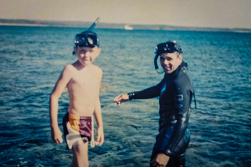 A young man in wetsuit and snorkelling gear stands in the water beside a young boy in swimming shorts and snorkel