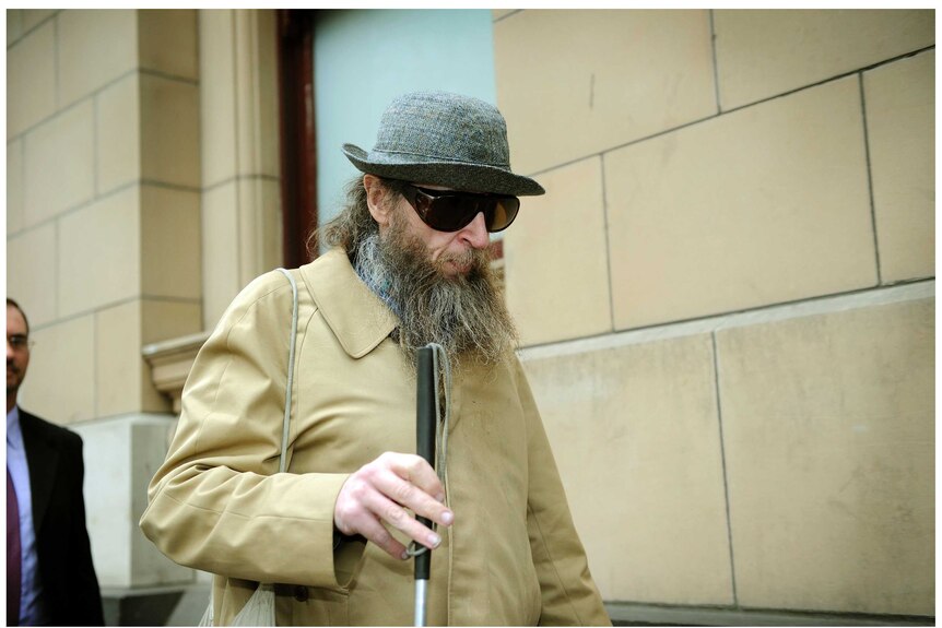 Robin Fletcher with a large beard, wearing a hat, sunglasses and carrying a cane, outside court.