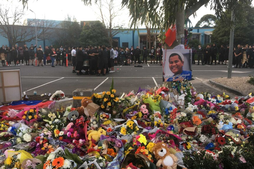 A sea of flowers lays in tribute near a photo of 15-year-old Solomone Taufeulungaki, with people dressed in black behind it.