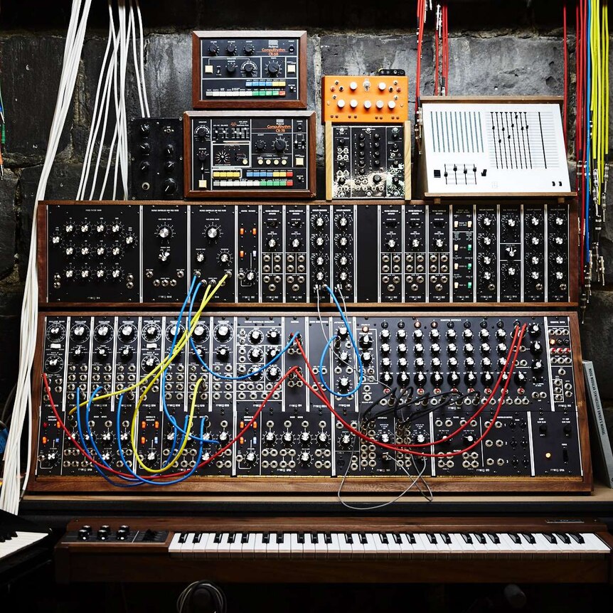 synthesiser keyboard with electronic equipment stacked on top covered with knobs and patch cords