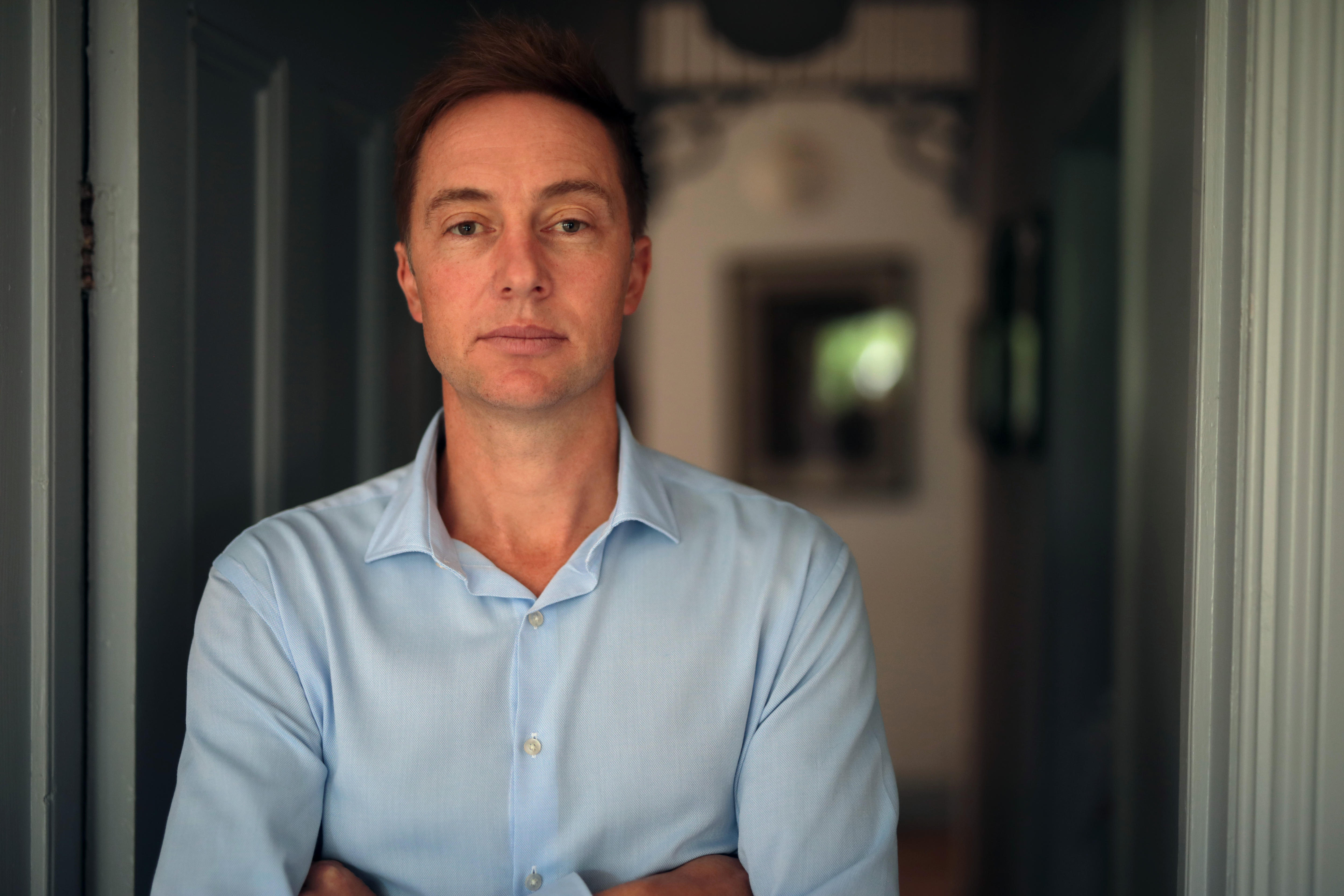 A man with his arms crossed stands at the door of a house. He is looking at the camera with a neutral expression