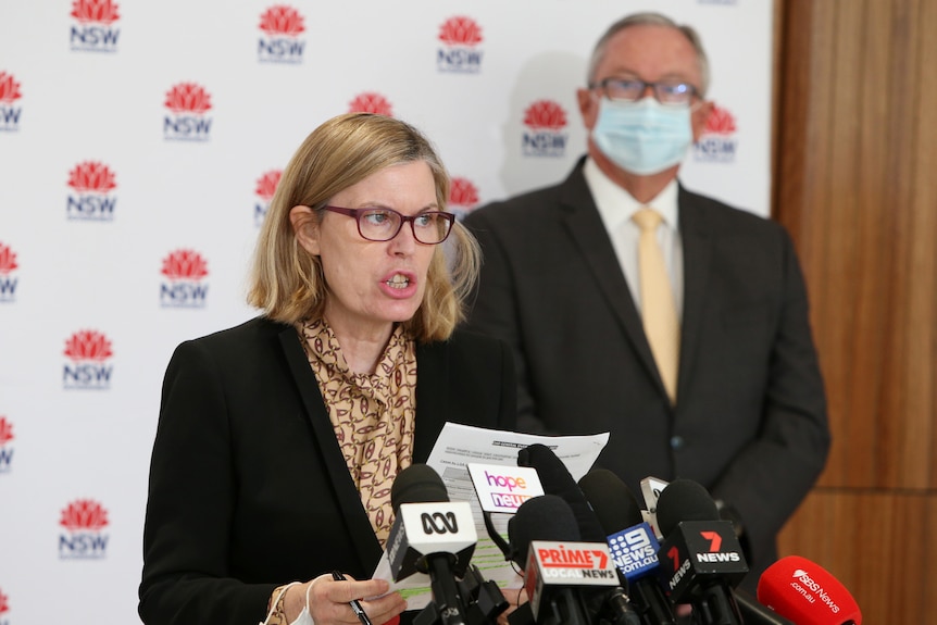 Blonde haired woman with red glasses and black jacket speaks to media