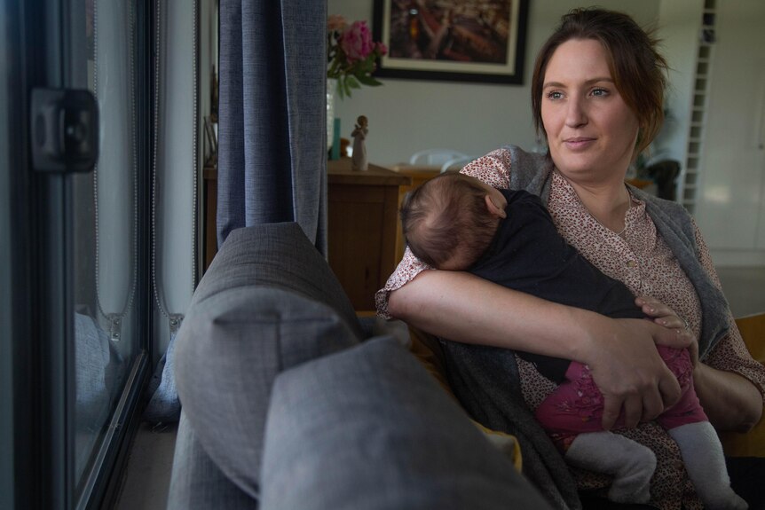 A serious-looking woman holding a baby sitting on a couch in a lounge room and looking out the window. 