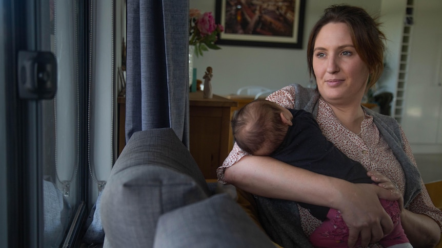 A serious-looking woman holding a baby sitting on a couch in a lounge room and looking out the window. 