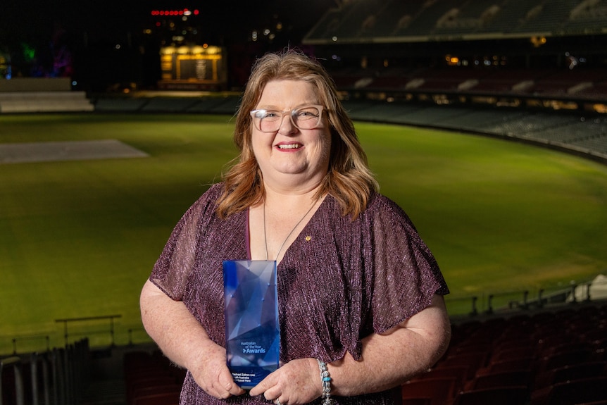 A woman in a purple formal dress holding a glass award in the stands of an oval