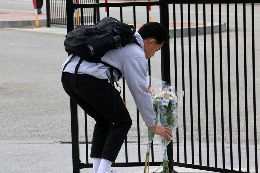 A man bends over at a metal gate, and sets down a bunch of white flowers.