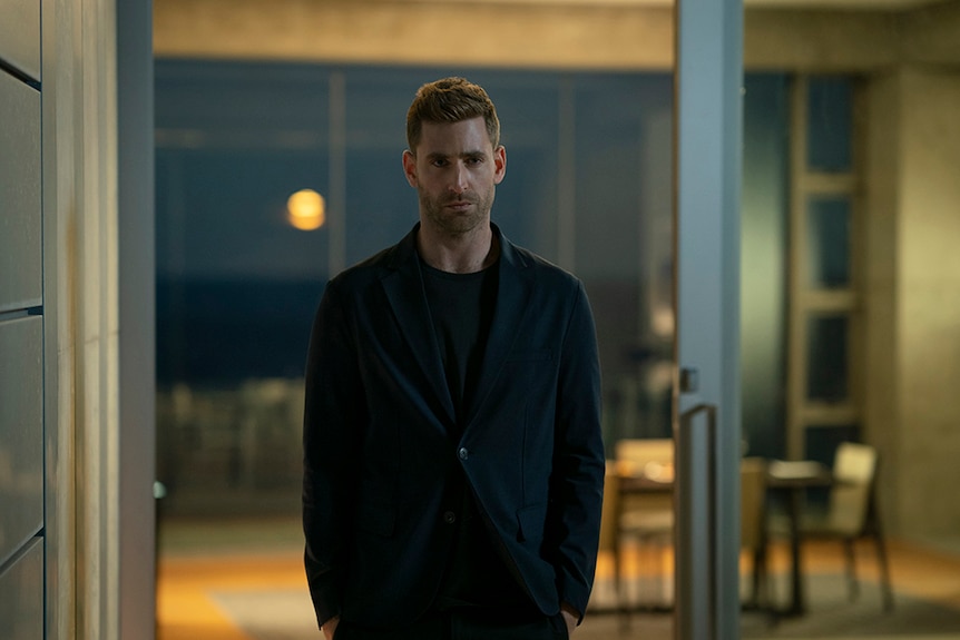 A man with short cropped hair and serious expression stands in black blazer and pants near sliding door in house at night.