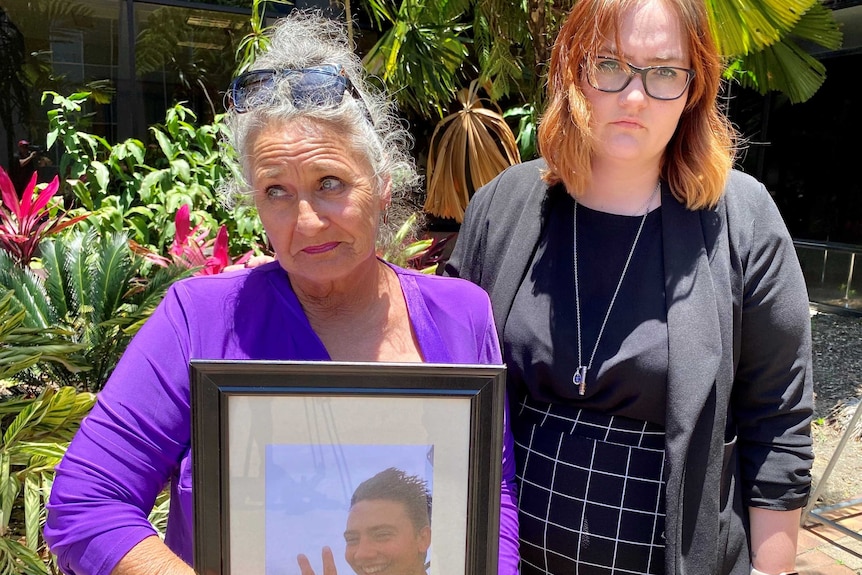 Two women look at the camera. One is holding a picture frame of her dead son.