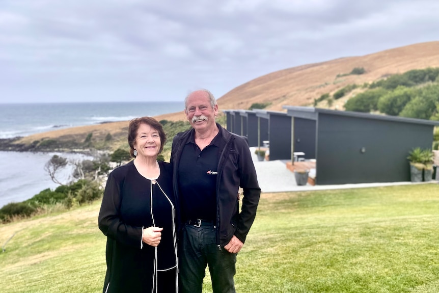 Kim and Bruce stand arm in arm in front of some of their cabins and the ocean