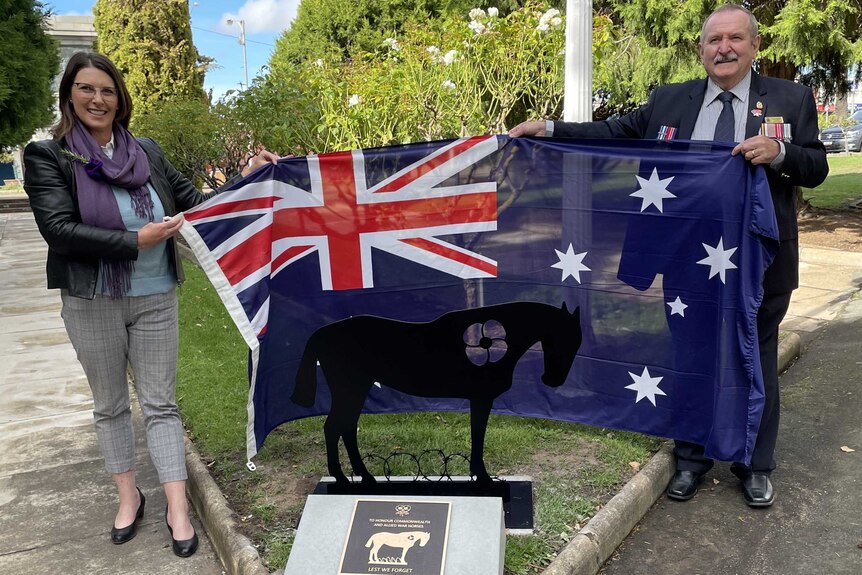 Two people hold an Australian flag behind a small steel sculpture of a horse in a garden.