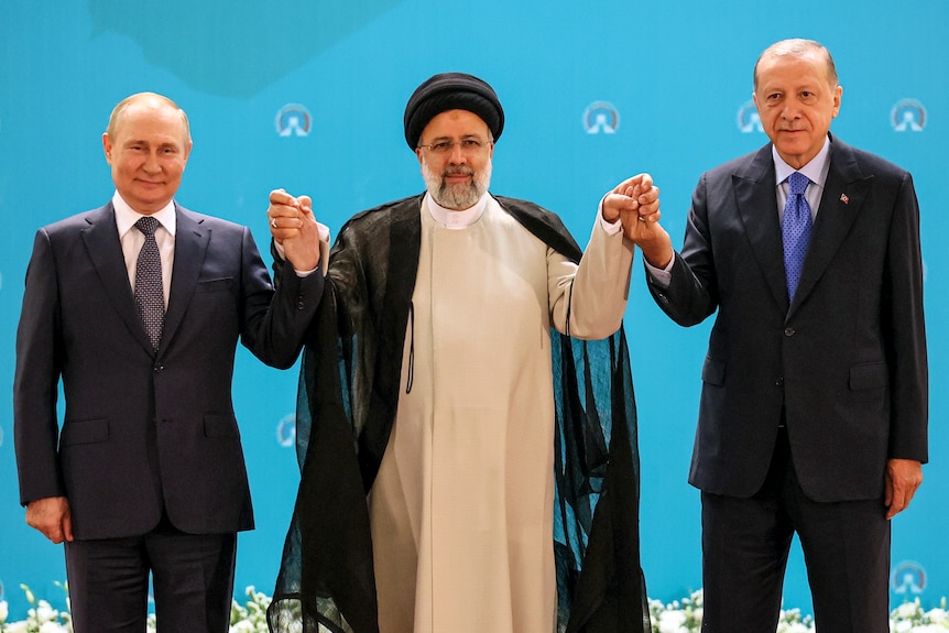 Two men in suits and one man in traditional Middle Eastern wear hold hands.