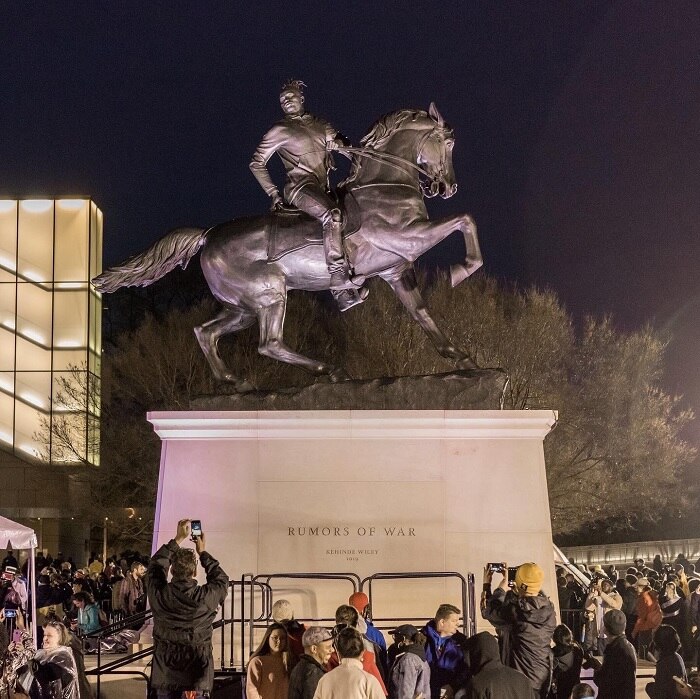 A crowd gathered is gathered around the Rumours of War statue, which shows an African-American man riding a horse.