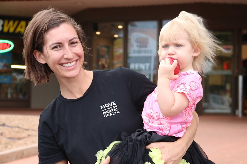 A young mother holding a small girl eating an apple in front of a supermarket.