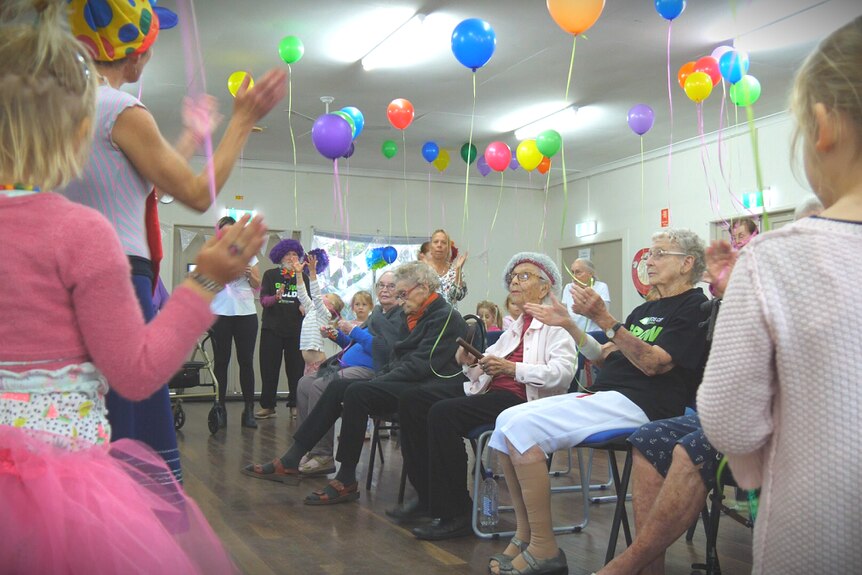 A group of elderly people sit on chairs clapping with young children around them and colourful balloons on the roof.
