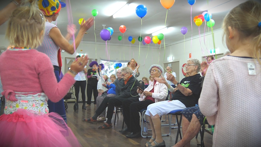 A group of elderly people sit on chairs clapping with young children around them and colourful balloons on the roof.