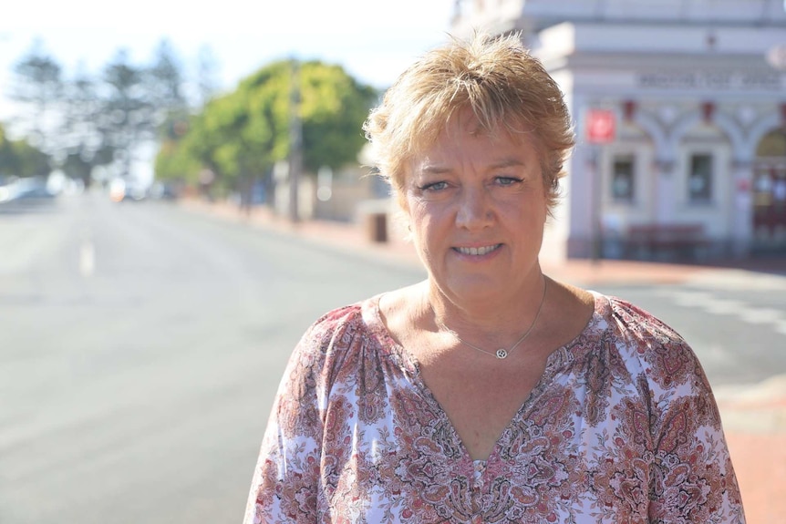 A blonde, smiling woman stands on a street corner wearing a red and white patterned top.