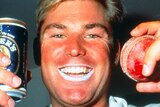 A smiling Shane Warne holds up a can of Foster's beer and a cricket ball