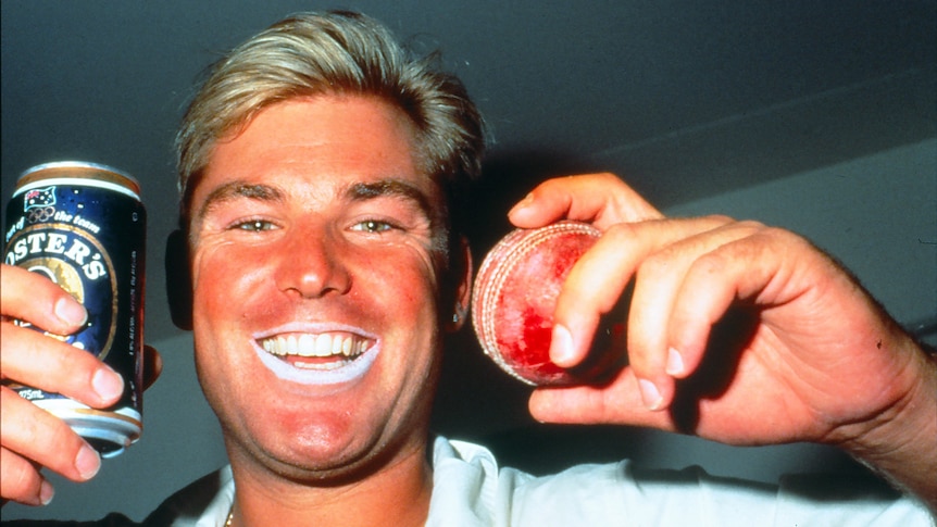 A smiling Shane Warne holds up a can of Foster's beer and a cricket ball