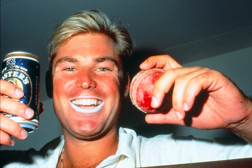 A smiling Shane Warne holds up a can of Foster's beer and a cricket ball