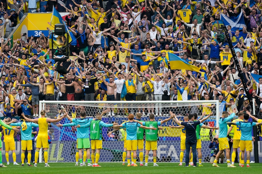 Ukrainian footballers stand in a line facing the stands with arms raised, as their fans wave flags and celebrate a win.