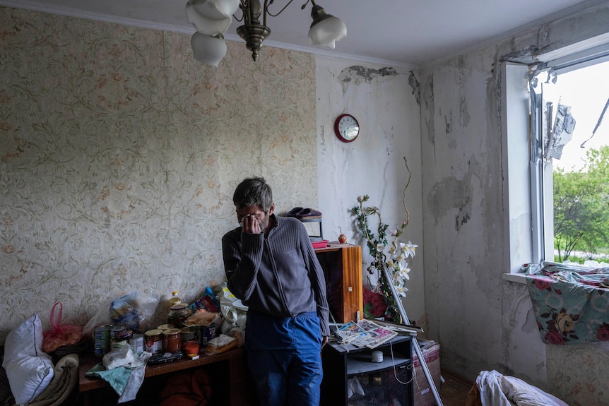 A man standing in a room of a house damaged by shelling wipes away tears. 