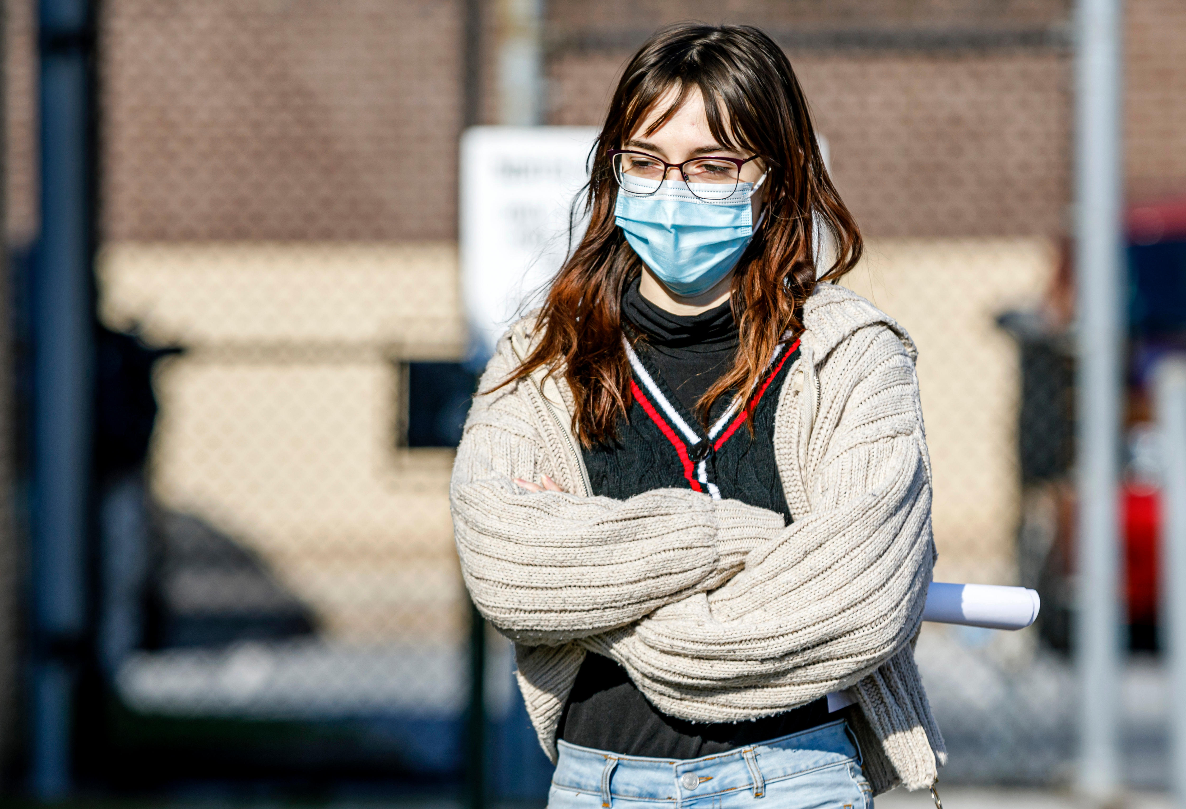 A brunette woman is pictured wearing glasses and a face mask. Her arms are crossed. She wears a white wool vest.
