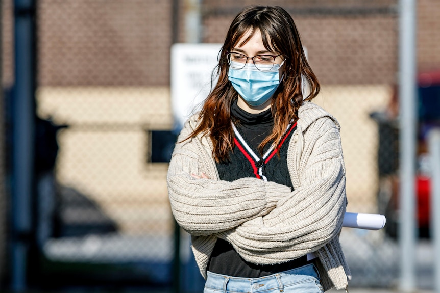 A brunette woman is pictured wearing glasses and a face mask. Her arms are crossed. She wears a white wool vest.