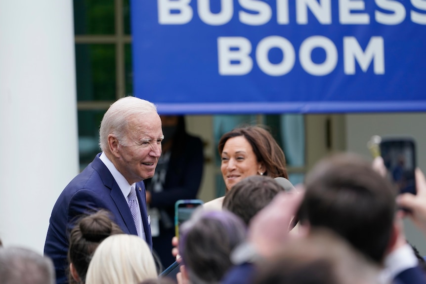 An image of Joe Biden surrounded by people, behind him is a blue banner with white letters. Next to him is Kamala Harris.
