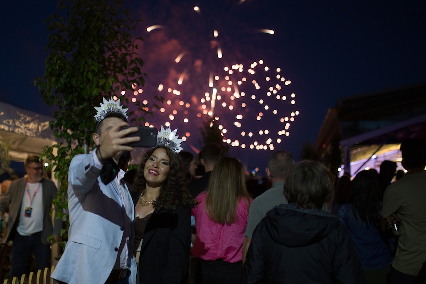 Two people wearing crowns and taking a selfie in front of fireworks