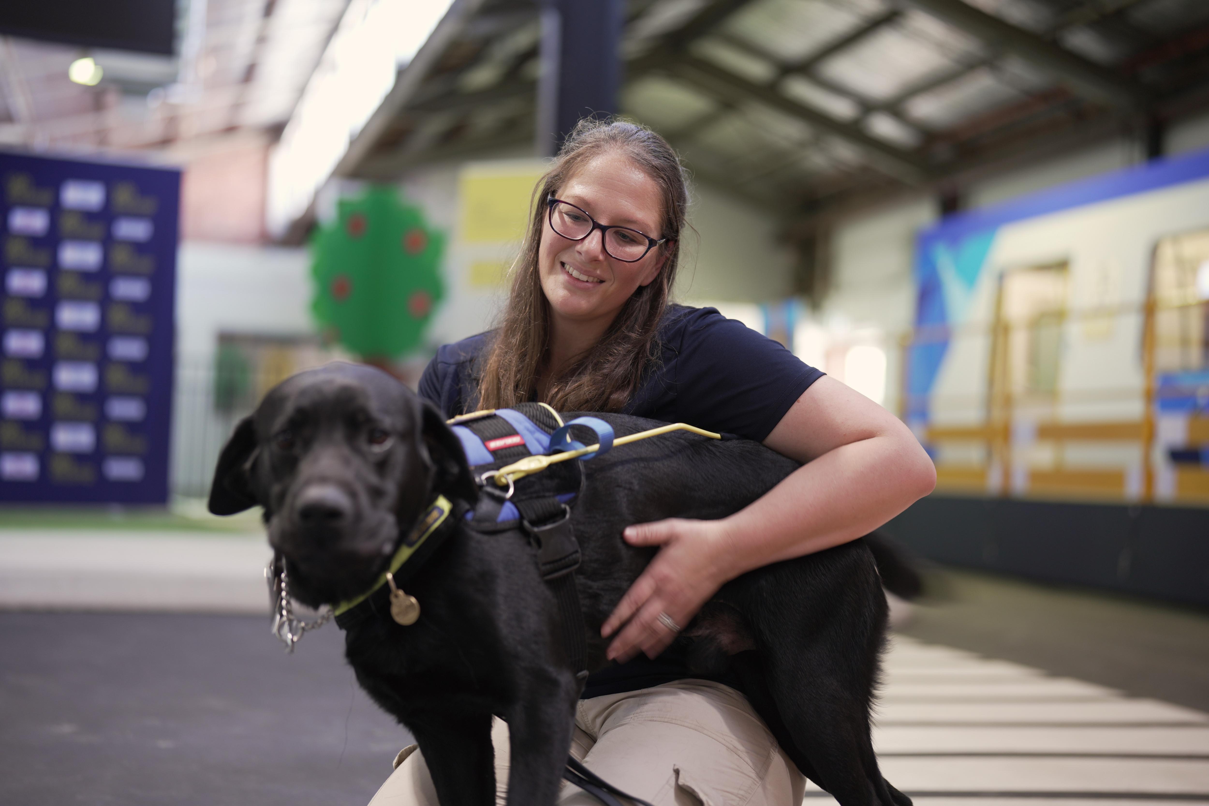 Brooke Anderson sonríe, se arrodilla y acaricia a un perro negro con arnés.
