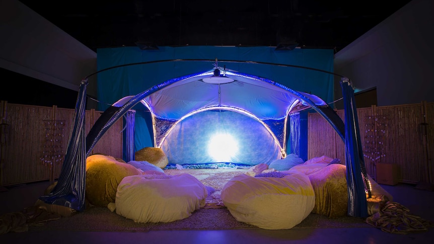 A tent like structure, lit but a filtered blue light, covers a carpeted area covered in gold beanbags.