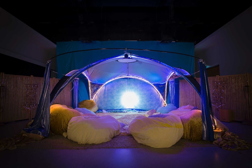 A tent like structure, lit but a filtered blue light, covers a carpeted area covered in gold beanbags.