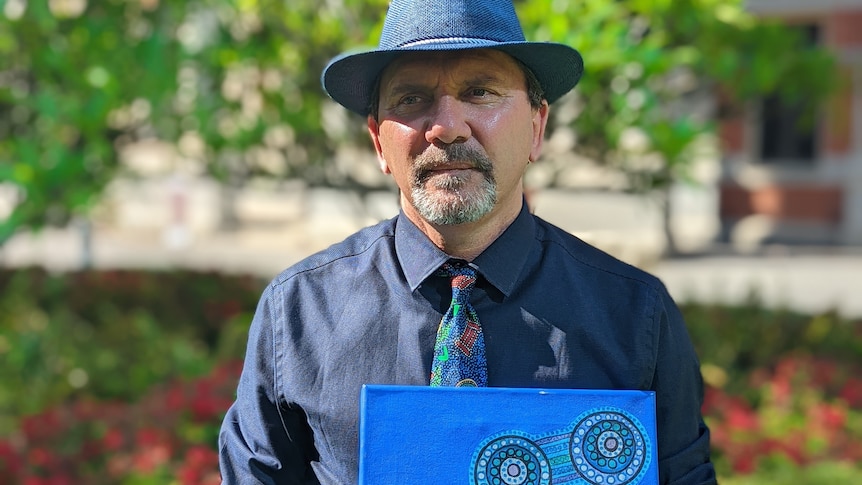 Man in fedora and tie stands outside holding blue dot painting by his missing sister.