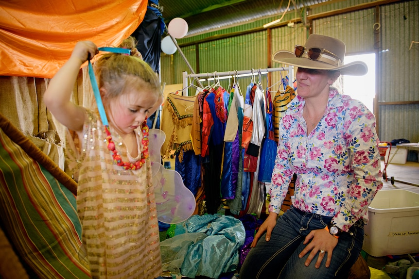A little girl putting a necklace over her head while a woman wearing a cowboy hat watches, inside a large shed. 