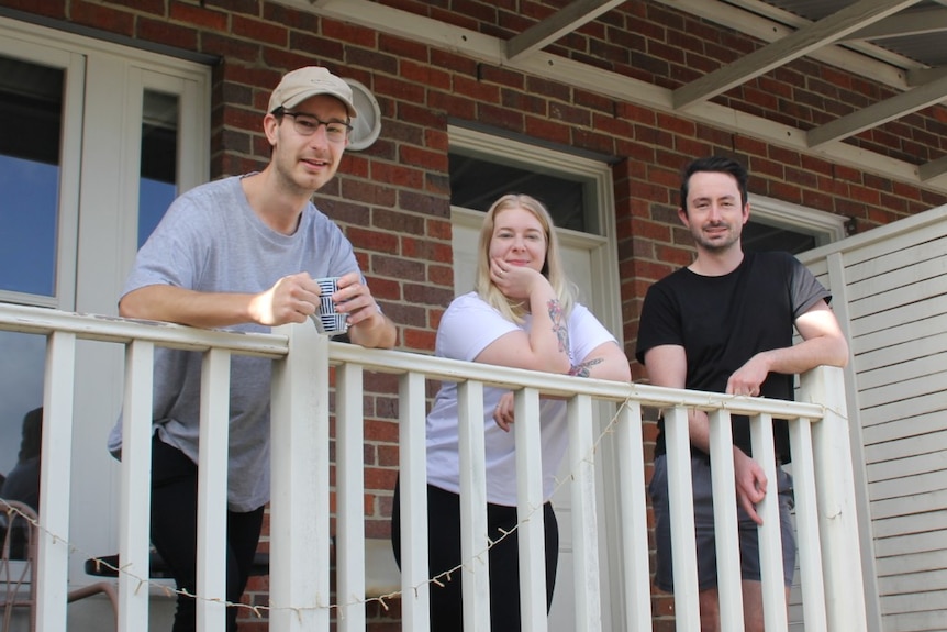 Two young man and a young woman lean over the railing in front of their home.