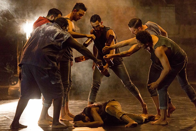 A group of male dancers surround and pour liquid from beer bottles onto a person huddled on ground in a hazy theatre.