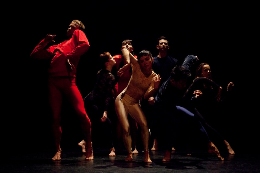 Seven dancers in matching long-sleeved shirts and fitted lycra pants contort their bodies on a darkened stage.