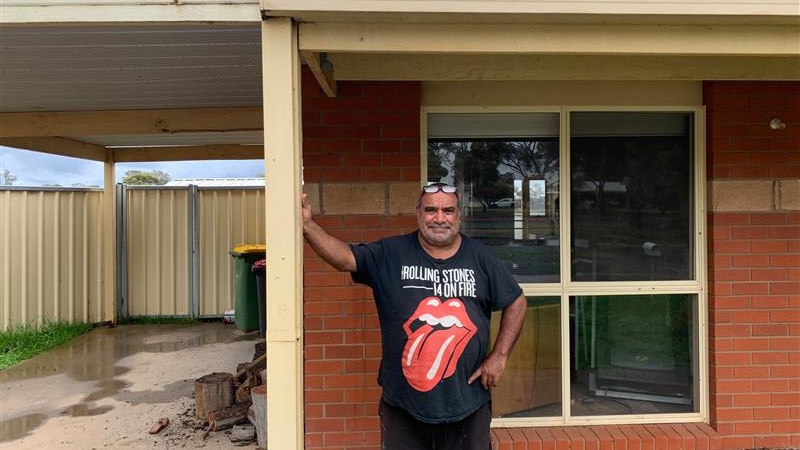Man leans on post at the front of his house, with a stern expression and a Rolling Stones t-shirt on