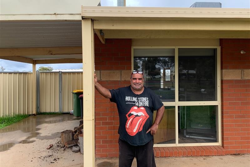 Man leans on post at the front of his house, with a stern expression and a Rolling Stones t-shirt on