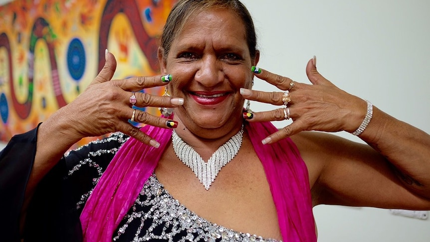 Aboriginal Woman Joyce Bonner, smiling into the camera, her hands are framing her face. An Indigenous artwork is behind her.
