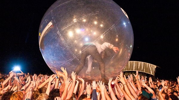 Wayne Coyne stands in a large inflatable dome on top of a large crowd of people who hold him up.