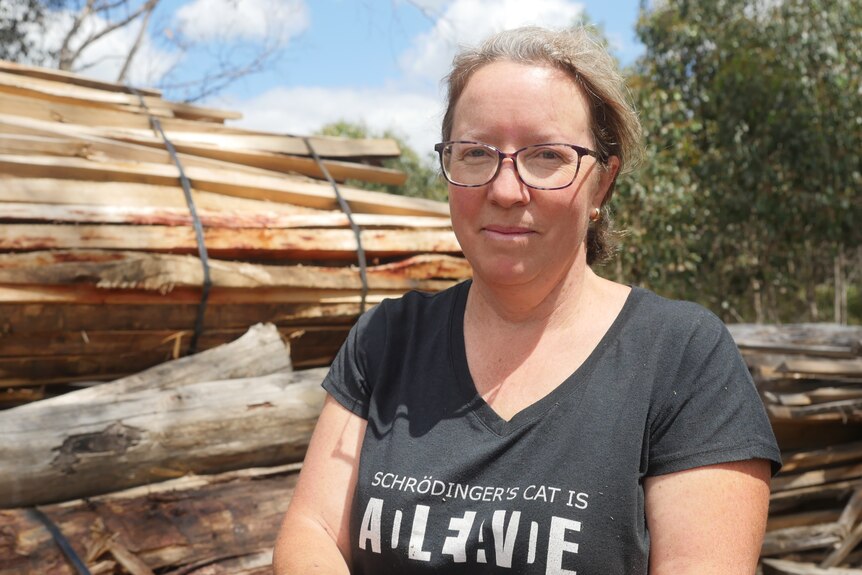 Woman with glasses stands in front of a large pile of cut and bundled timber. 
