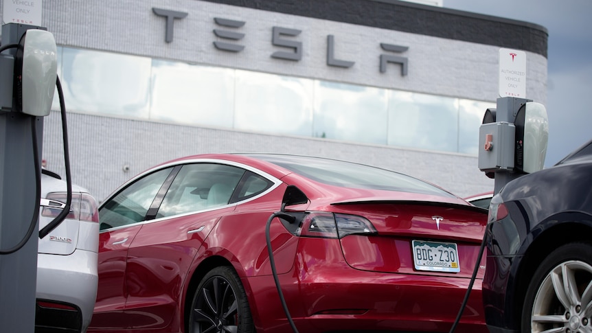 a red Tesla Model 3 sedan is charged at a Tesla dealership with the Tesla logo shown on the dealership building in the back