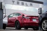 a red Tesla Model 3 sedan is charged at a Tesla dealership with the Tesla logo shown on the dealership building in the back