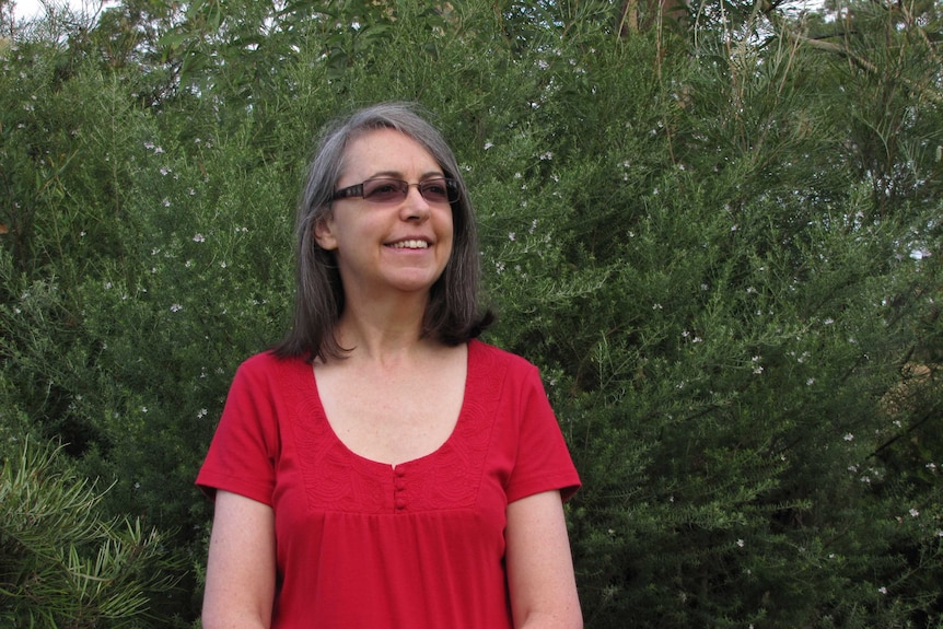 A woman with grey hair wearing a red shirt stands in front of a bush.